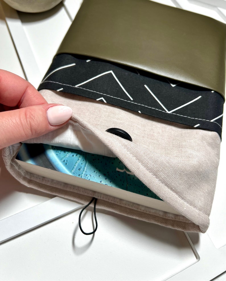 Pu&ograve; includere: Una copertina per libro con uno strato esterno in lino beige, uno strato di tessuto geometrico bianco e nero e una patta in pelle verde scuro. La copertina ha un bottone nero e un segnalibro elastico nero.