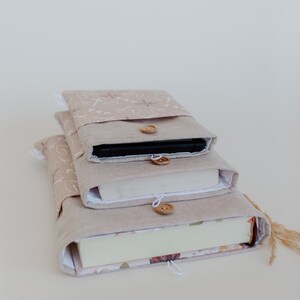 Puede incluir: Tres fundas de libros beige apiladas. La funda superior contiene un tel&eacute;fono, la del medio un libro y la inferior un libro con estampado floral. Cada funda tiene un cierre de bot&oacute;n de madera.