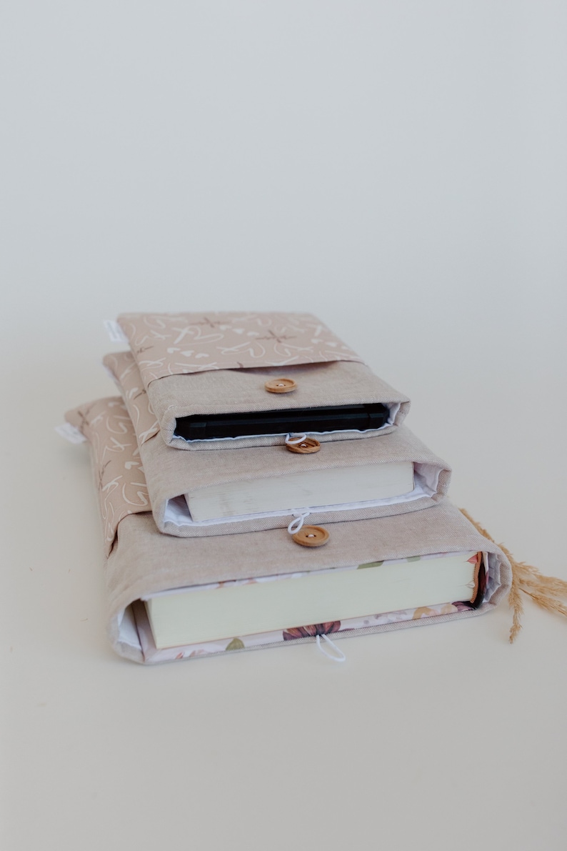 Puede incluir: Tres fundas de libros beige apiladas. La funda superior contiene un tel&eacute;fono, la del medio un libro y la inferior un libro con estampado floral. Cada funda tiene un cierre de bot&oacute;n de madera.