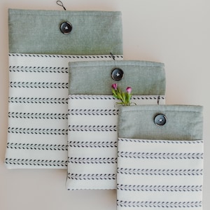 May include: Three fabric pouches in varying sizes. Each pouch has a sage green top and a white body with a repeating black leaf pattern. Black buttons secure the tops. One pouch holds small pink flowers.