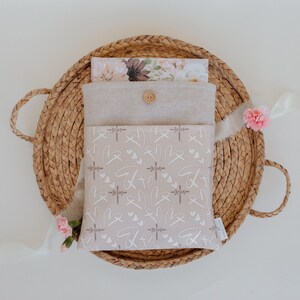 Puede incluir: Una funda de tela beige con cierre de bot&oacute;n, con un patr&oacute;n de cruces, corazones y la palabra "Jes&uacute;s" en blanco. La funda est&aacute; colocada dentro de una cesta tejida, con una tarjeta con estampado floral y flores rosas.