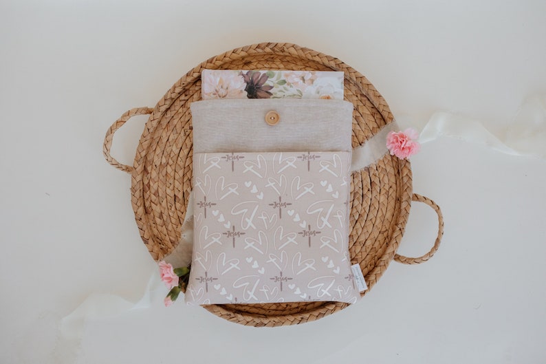 Puede incluir: Una funda de tela beige con cierre de bot&oacute;n, con un patr&oacute;n de cruces, corazones y la palabra "Jes&uacute;s" en blanco. La funda est&aacute; colocada dentro de una cesta tejida, con una tarjeta con estampado floral y flores rosas.