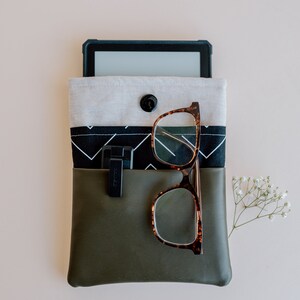 Pu&ograve; includere: Una pochette verde con cerniera con una fodera in tessuto a motivi bianchi e neri. Un e-reader nero e un paio di occhiali marroni sono all'interno della pochette.