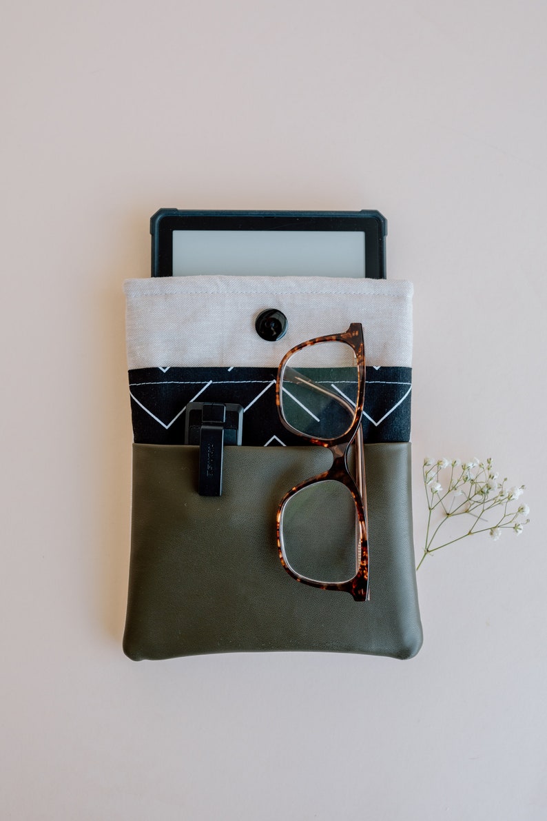 Pu&ograve; includere: Una pochette verde con cerniera con una fodera in tessuto a motivi bianchi e neri. Un e-reader nero e un paio di occhiali marroni sono all'interno della pochette.