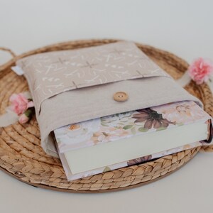 Puede incluir: Una funda de libro beige con cierre de bot&oacute;n, que contiene un libro con arte de portada floral. La funda est&aacute; sobre una bandeja tejida con flores rosas. La funda tiene un patr&oacute;n claro.