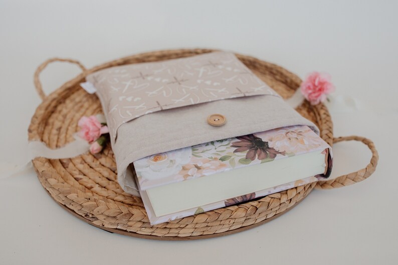 Puede incluir: Una funda de libro beige con cierre de bot&oacute;n, que contiene un libro con arte de portada floral. La funda est&aacute; sobre una bandeja tejida con flores rosas. La funda tiene un patr&oacute;n claro.