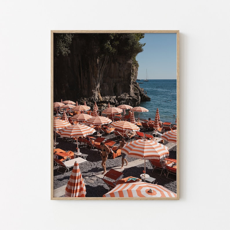 May include: A beach scene with orange and white striped beach umbrellas and lounge chairs. Two people are walking on the beach in front of the umbrellas. The beach is located near a rocky cliff and the ocean is in the background.