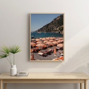 May include: A framed photograph of a beach scene with orange and white striped beach umbrellas and lounge chairs on a pebble beach. The beach is located in front of a rocky cliff and a blue ocean. There are boats in the distance.