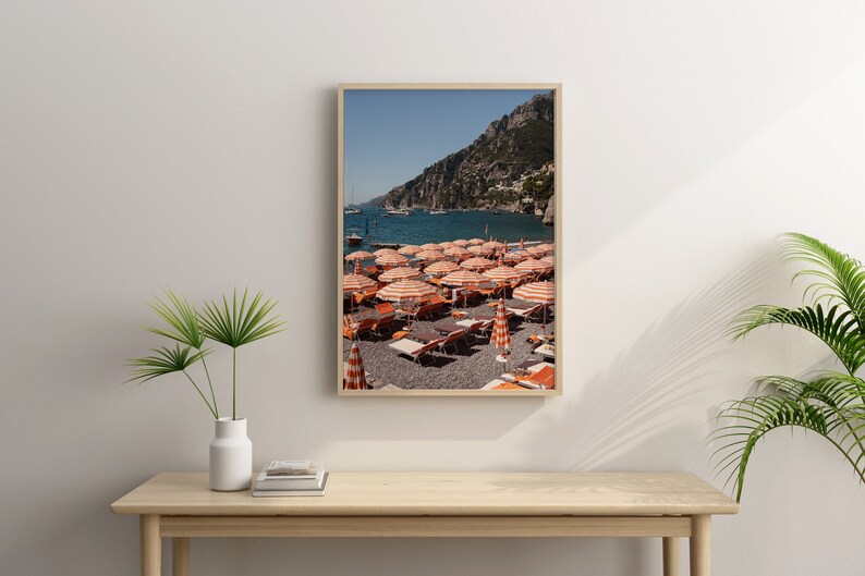 May include: A framed photograph of a beach scene with orange and white striped beach umbrellas and lounge chairs on a pebble beach. The beach is located in front of a rocky cliff and a blue ocean. There are boats in the distance.