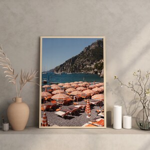 May include: A framed photograph of a beach scene with orange and white striped beach umbrellas and lounge chairs on a pebble beach. The beach is located in front of a rocky cliff and a clear blue ocean.