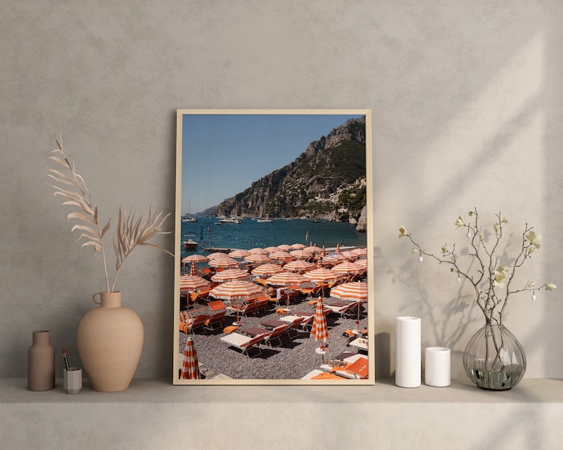 May include: A framed photograph of a beach scene with orange and white striped beach umbrellas and lounge chairs on a pebble beach. The beach is located in front of a rocky cliff and a clear blue ocean.
