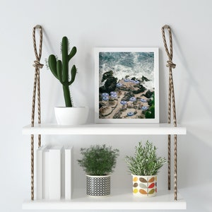 May include: White floating shelves with rope hangers, featuring a cactus, two potted plants, a framed photograph of a beach scene, and a stack of books.