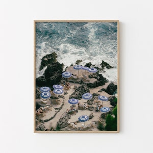 May include: An aerial view of a rocky beach with blue and white striped umbrellas and chairs. The ocean waves are crashing against the rocks in the background.