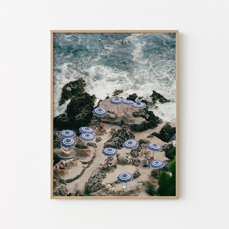 May include: An aerial view of a rocky beach with blue and white striped umbrellas and chairs. The ocean waves are crashing against the rocks in the background.