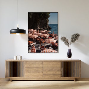 May include: A framed photograph of a beach scene with red and white striped umbrellas and people walking on the sand. The beach is surrounded by cliffs and the ocean is in the background.