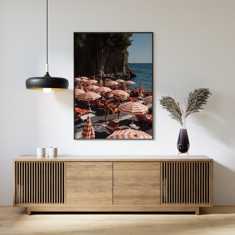 May include: A framed photograph of a beach scene with red and white striped umbrellas and people walking on the sand. The beach is surrounded by cliffs and the ocean is in the background.