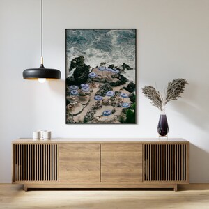 May include: A framed photograph of a beach scene with blue and white striped umbrellas and people relaxing on the sand. The photo is hanging on a white wall above a wooden cabinet.