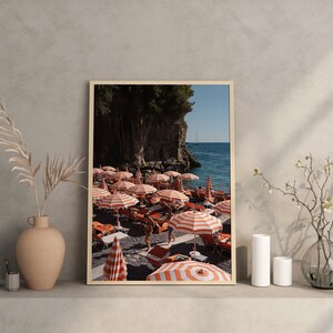 May include: A framed photograph of a beach scene with red and white striped beach umbrellas and people relaxing on the sand. The beach is located on a rocky coastline with a clear blue ocean in the background.