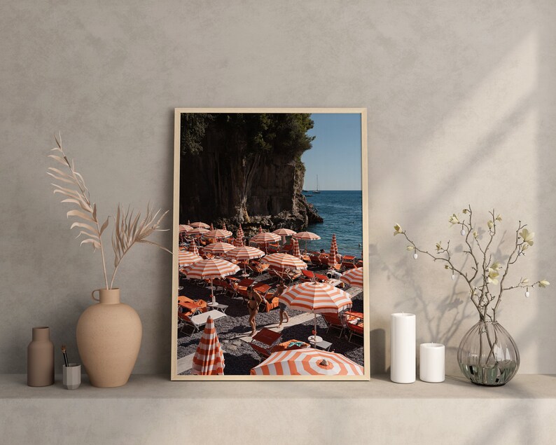 May include: A framed photograph of a beach scene with red and white striped beach umbrellas and people relaxing on the sand. The beach is located on a rocky coastline with a clear blue ocean in the background.