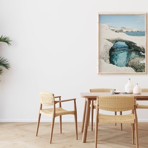 May include: A dining room with a wooden table and chairs. The chairs are woven with a light brown material. A framed photograph of a rock formation with a blue lagoon hangs on the wall. A potted palm tree is in the corner of the room.