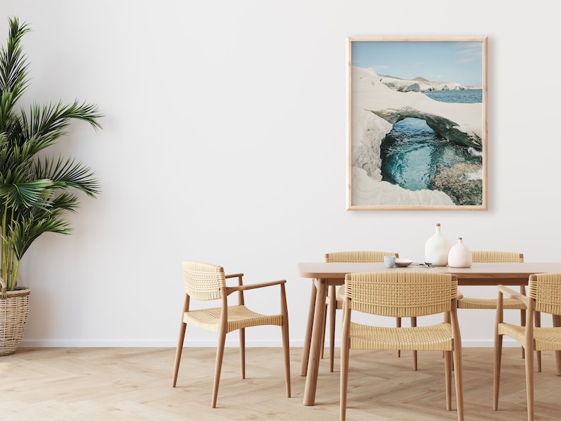 May include: A dining room with a wooden table and chairs. The chairs are woven with a light brown material. A framed photograph of a rock formation with a blue lagoon hangs on the wall. A potted palm tree is in the corner of the room.