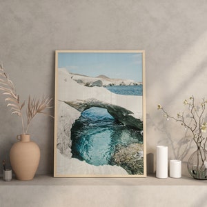 May include: A framed photograph of a natural rock archway over a turquoise ocean. The archway is made of white rock and the ocean is a bright blue. The photo is set on a shelf with a vase of dried branches and two white ceramic vases.