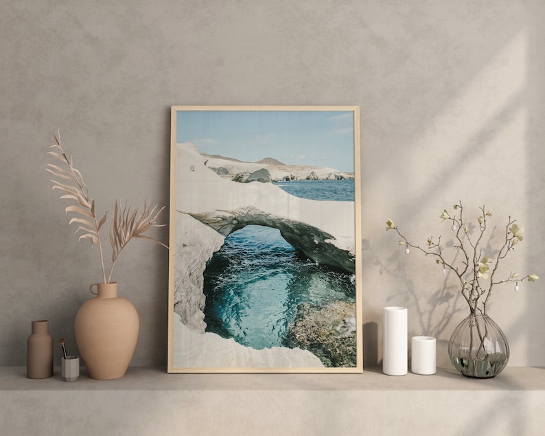 May include: A framed photograph of a natural rock archway over a turquoise ocean. The archway is made of white rock and the ocean is a bright blue. The photo is set on a shelf with a vase of dried branches and two white ceramic vases.