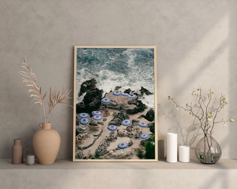 May include: A framed photograph of a rocky coastline with blue and white striped umbrellas and chairs. The water is crashing against the rocks and there are people sitting under the umbrellas.