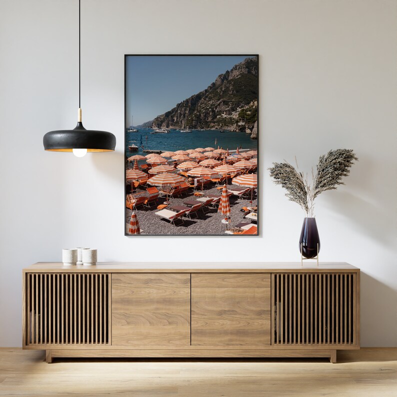 May include: A framed photograph of a beach scene with orange and white striped beach umbrellas and lounge chairs on a pebbled beach. The beach is located in a bay with a rocky cliff in the background.