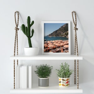 May include: White floating shelves with rope hangers, featuring a framed photograph of a beach scene with colorful umbrellas, a potted cactus, and two potted plants. The shelves are decorated with books and decorative pots.