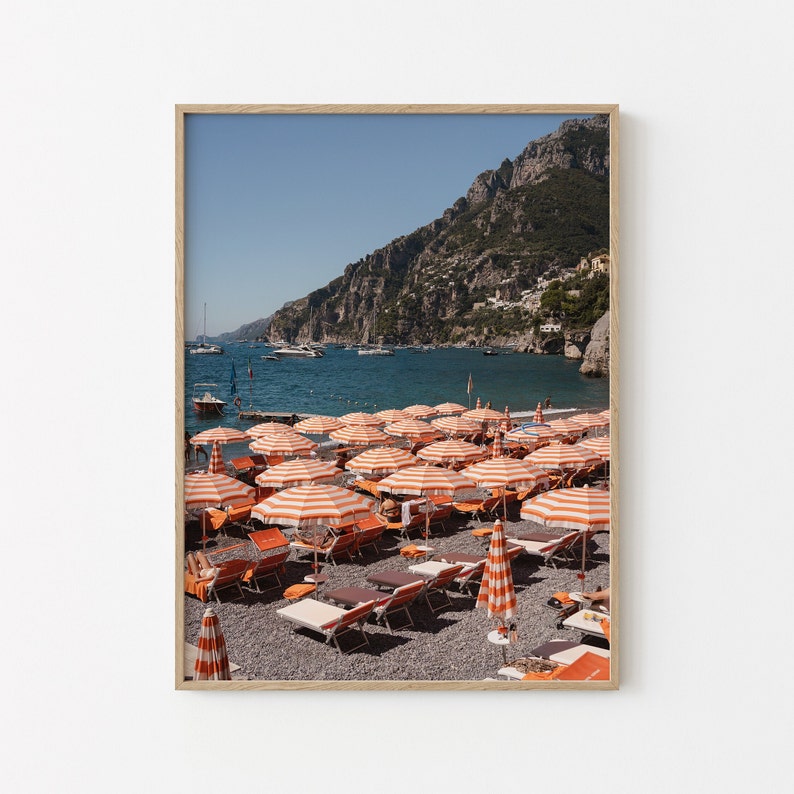 May include: A beach scene with orange and white striped beach umbrellas and lounge chairs on a pebble beach. The beach is located in front of a rocky cliffside with a clear blue ocean in the background.