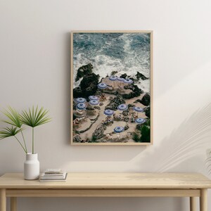 May include: An aerial view of a rocky beach with blue and white striped umbrellas and people relaxing on the sand. The ocean waves are crashing against the rocks in the background.