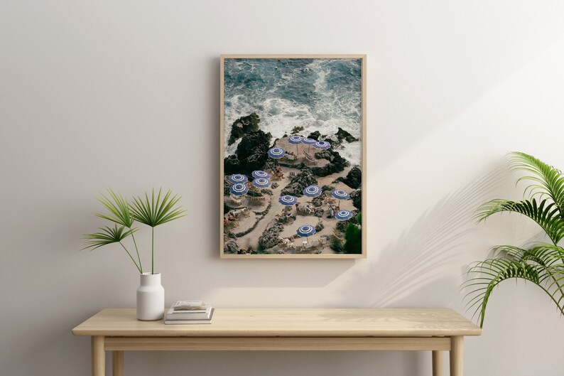 May include: An aerial view of a rocky beach with blue and white striped umbrellas and people relaxing on the sand. The ocean waves are crashing against the rocks in the background.