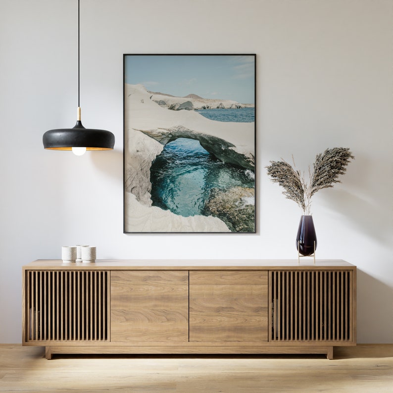 May include: A framed photograph of a natural rock archway over a turquoise ocean. The photo is hanging above a wooden cabinet with two doors and slatted panels.