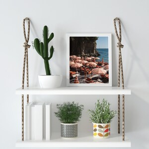May include: White floating shelves with rope hangers, featuring a framed photograph of a beach scene with colorful umbrellas, a green cactus in a white pot, a potted plant with black and white patterned pot, and a potted plant with a colorful leaf patterned pot.