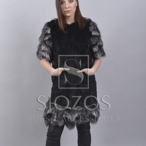 May include: A black fur coat with silver fox trim on the sleeves and hem. The coat is knee-length and has a simple, elegant design. The model is wearing black trousers and boots.