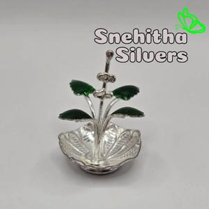 May include: A silver-colored decorative piece with a leaf design and green accents. The piece has a bowl-like base and a floral arrangement with green leaves. The text "Snehitha Silvers" is displayed above the object.