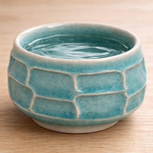 May include: A turquoise ceramic cup filled with water. The cup has a textured, geometric pattern and a white rim. The water's surface reflects light, and the cup sits on a light-colored wooden surface. The cup is a small, handmade item.
