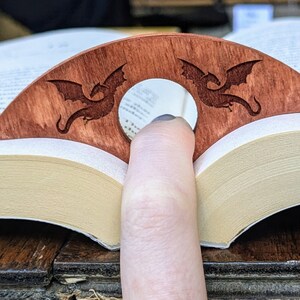 May include: A wooden bookmark with two dragon silhouettes carved into it. The bookmark is brown and has a hole in the center for a finger to hold it in place.
