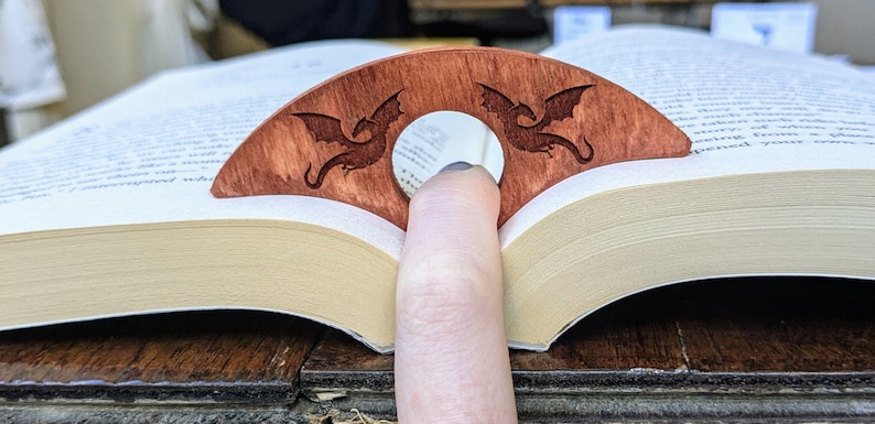 May include: A wooden bookmark with two dragon silhouettes carved into it. The bookmark is brown and has a hole in the center for a finger to hold it in place.