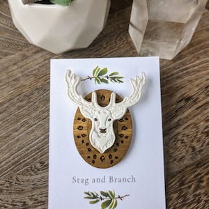 May include: White wooden stag head pin with a brown wooden oval background featuring a leaf design. The pin is displayed on a white card with the text "Stag and Branch".