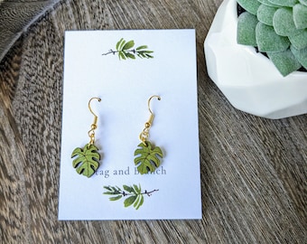 May include: A pair of green monstera leaf earrings with gold hooks. The earrings are displayed on a white card with the text "Tag and Branch".