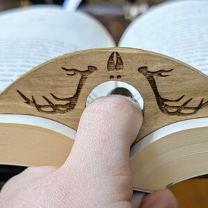 May include: A wooden book holder with a carved design of two deer antlers. The holder is shaped like a crescent moon and has a hole in the center for a finger to hold it in place.