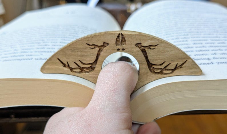 May include: A wooden book holder with a carved design of two deer antlers. The holder is shaped like a crescent moon and has a hole in the center for a finger to hold it in place.