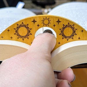 May include: A wooden bookmark shaped like a crescent moon with a hole in the center for a finger. The bookmark is decorated with a sun and stars design in a light brown color.