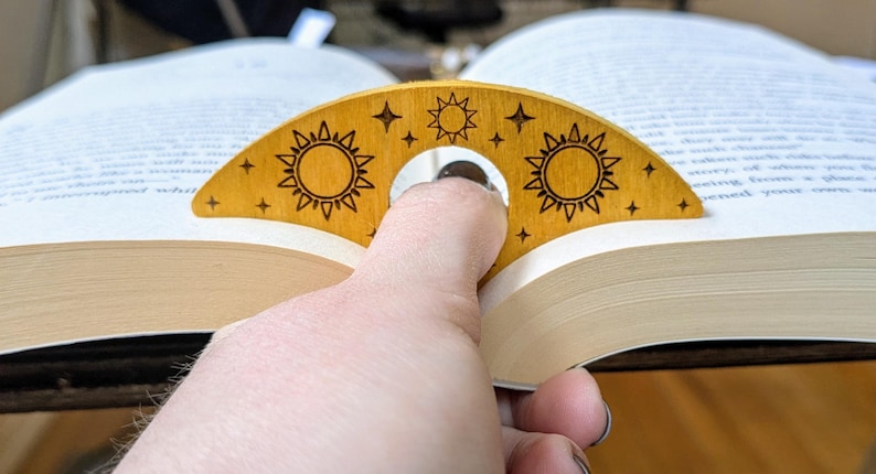 May include: A wooden bookmark shaped like a crescent moon with a hole in the center for a finger. The bookmark is decorated with a sun and stars design in a light brown color.