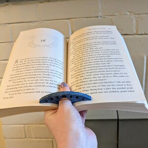 May include: A blue book with a blue crescent moon bookmark. The book is open to page 16 and the text reads "The Bone Witch" and "Actually, I was wondering if you could design something for my friend here."
