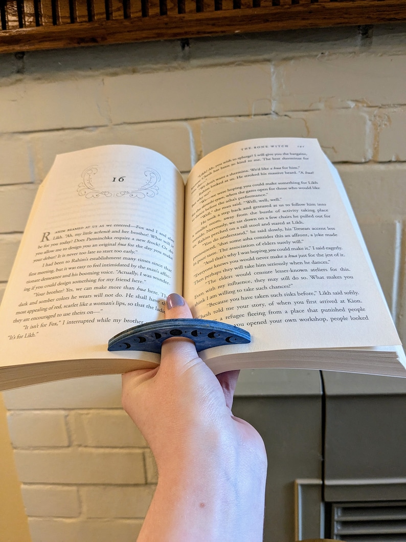 May include: A blue book with a blue crescent moon bookmark. The book is open to page 16 and the text reads "The Bone Witch" and "Actually, I was wondering if you could design something for my friend here."