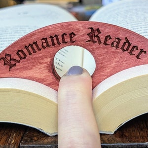 May include: A wooden bookmark shaped like a crescent moon with a mirror in the center. The bookmark is red and has the words "Romance Reader" engraved on it in a gothic font.
