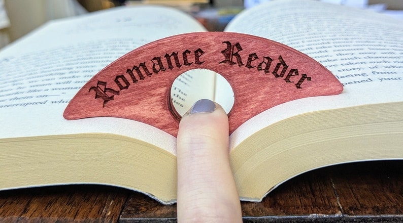 May include: A wooden bookmark shaped like a crescent moon with a mirror in the center. The bookmark is red and has the words "Romance Reader" engraved on it in a gothic font.
